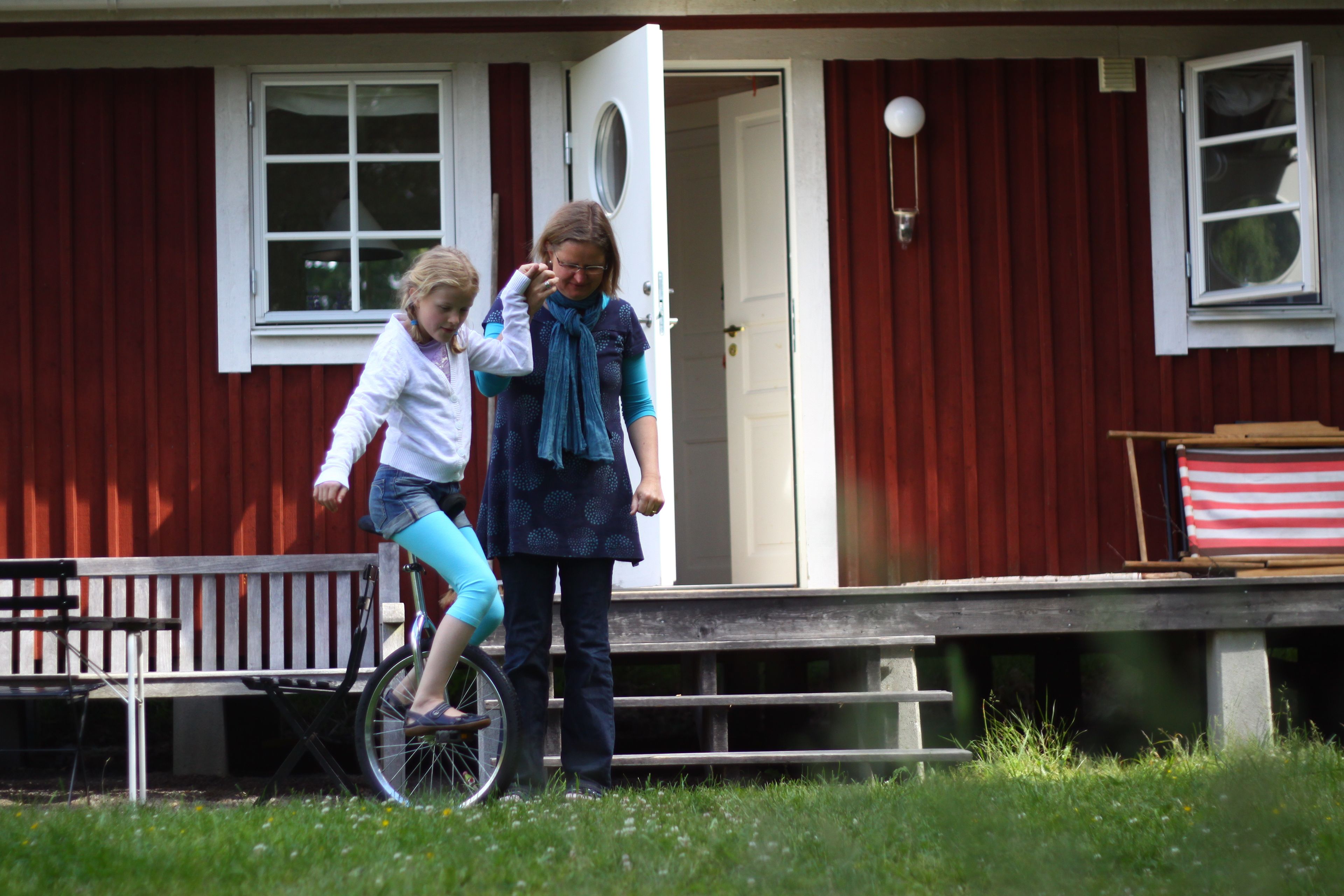 Ett hus med en veranda, ett barn på en cykel och en vuxen som står i närheten.