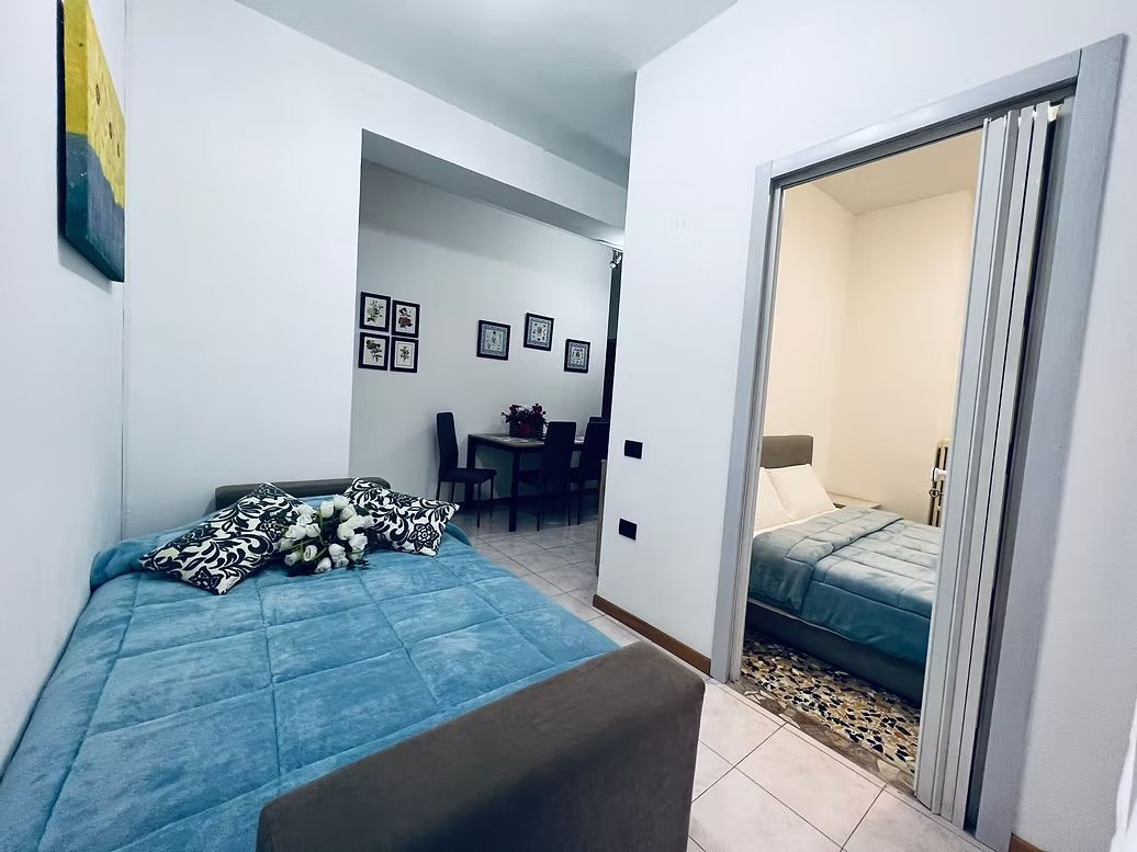 A small, modern room with a bed, a desk, and a chair. There are framed pictures on the wall and a door leading to another room.