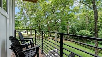 A wooden deck with Adirondack chairs, a metal railing, and a view of a lush green landscape with trees.