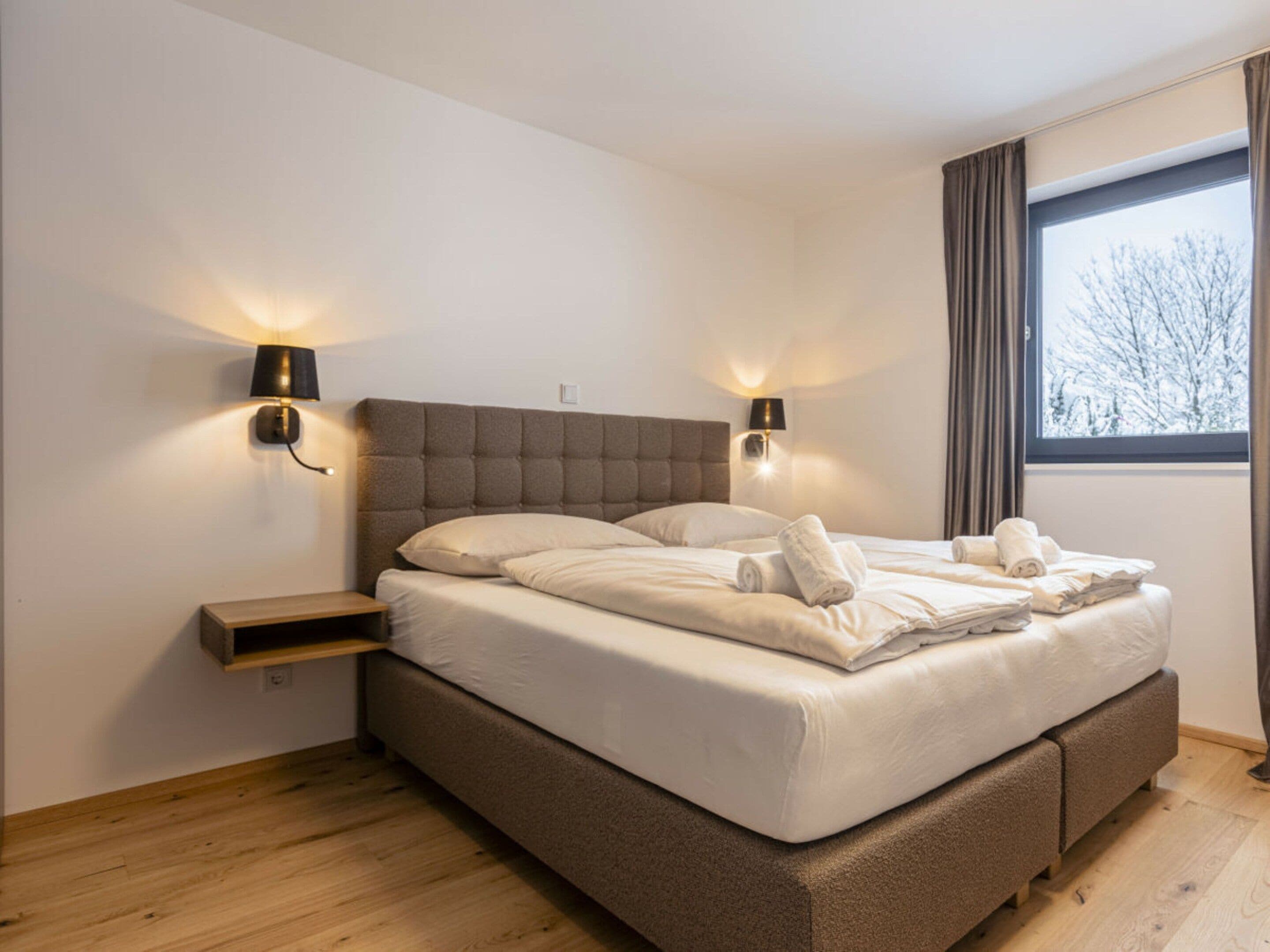 A modern bedroom with a large bed, two bedside lamps, a nightstand, and a window with curtains.
