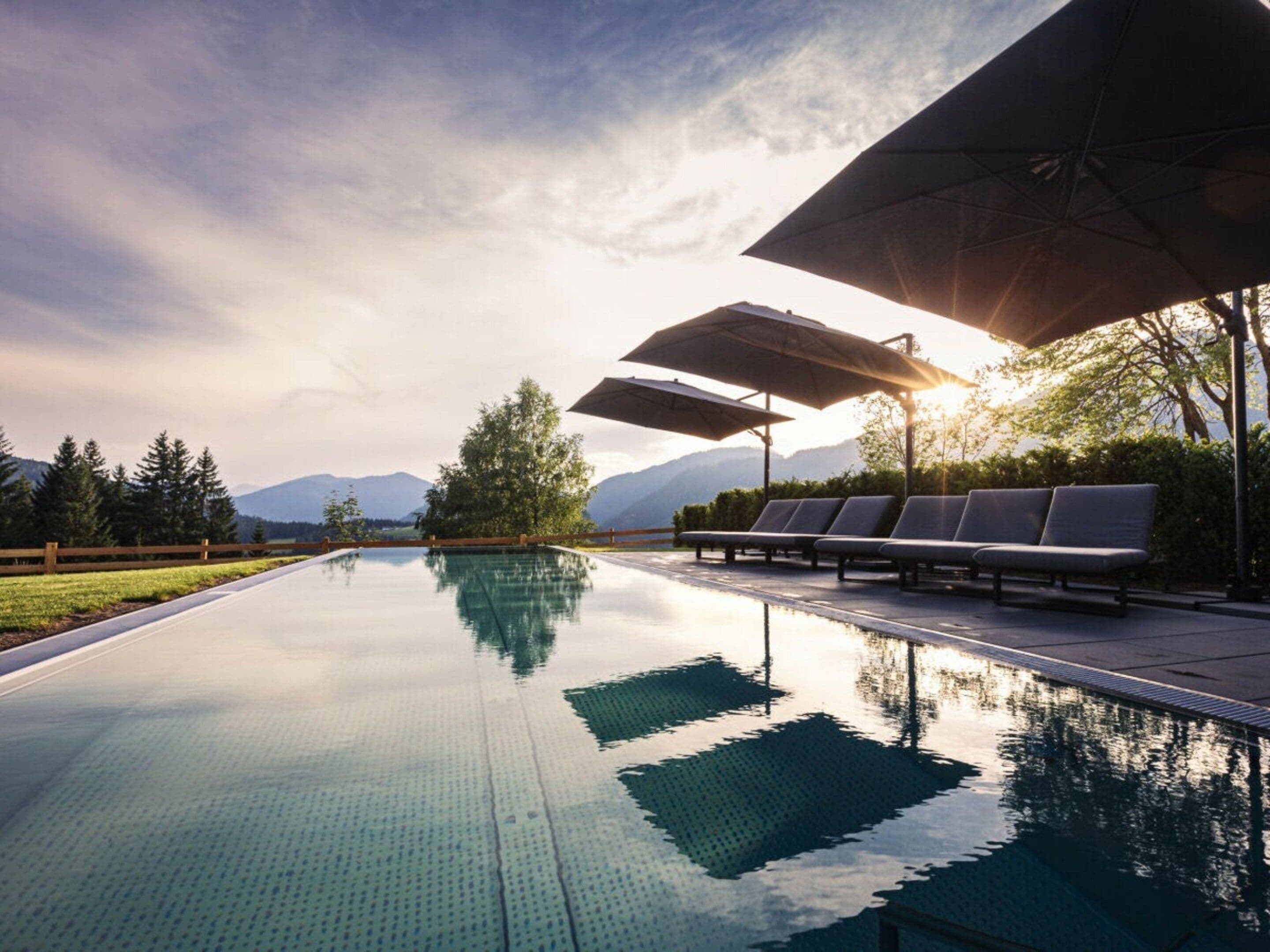 Ein Swimmingpool mit Liegestühlen und Sonnenschirmen, umgeben von Bäumen und Bergen im Hintergrund.