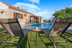Un área junto a la piscina con dos sillas de descanso, una mesita y un edificio de piedra al fondo.