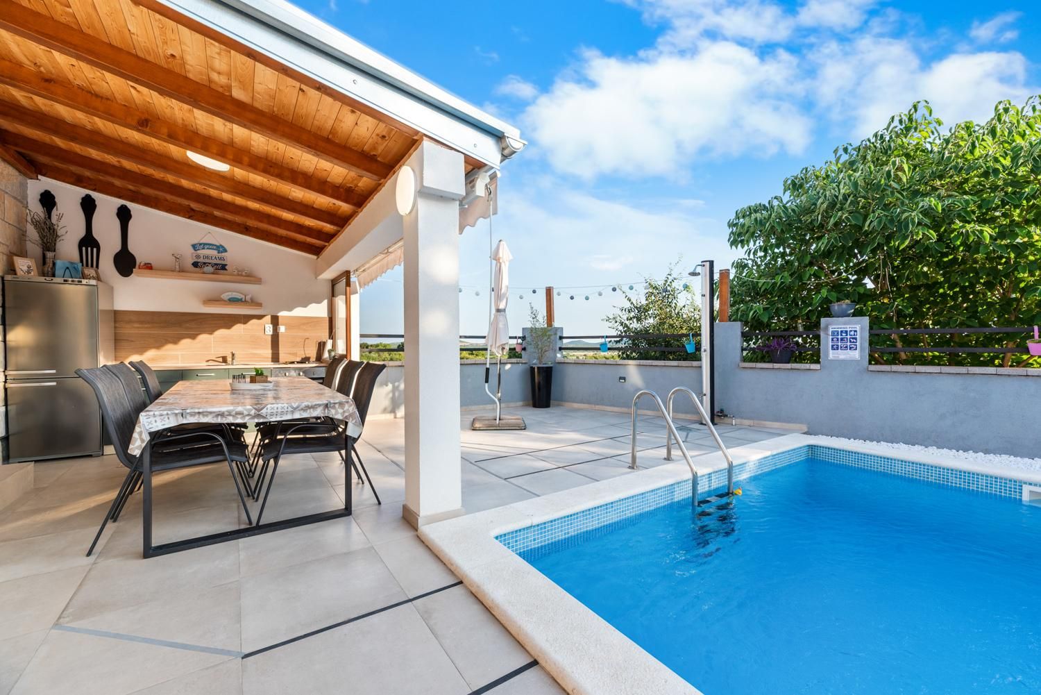 A covered outdoor area with a pool, dining table, and chairs, and a kitchen area with a refrigerator.