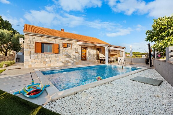 Una casa de piedra con techo de tejas, piscina y jardín con guijarros blancos.