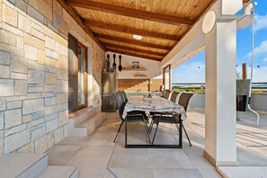 Un área de comedor al aire libre con pared de piedra, techo de madera y vista al mar.