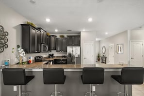 A modern kitchen with dark cabinets, stainless steel appliances, and a granite countertop. There are three black bar stools in front of the counter. - 9BR Disney Getaway with Themed Rooms and Theater (Four Corners)