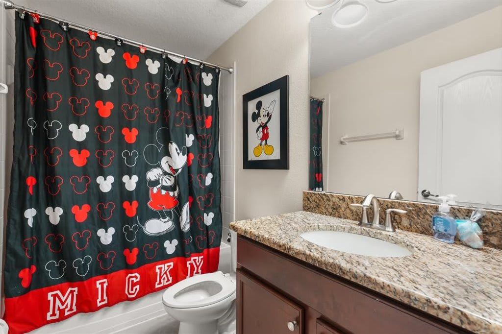 Un baño con cortina de ducha de Mickey Mouse, un cuadro enmarcado de Mickey en la pared, un inodoro blanco y una encimera de granito con un lavabo y grifo.