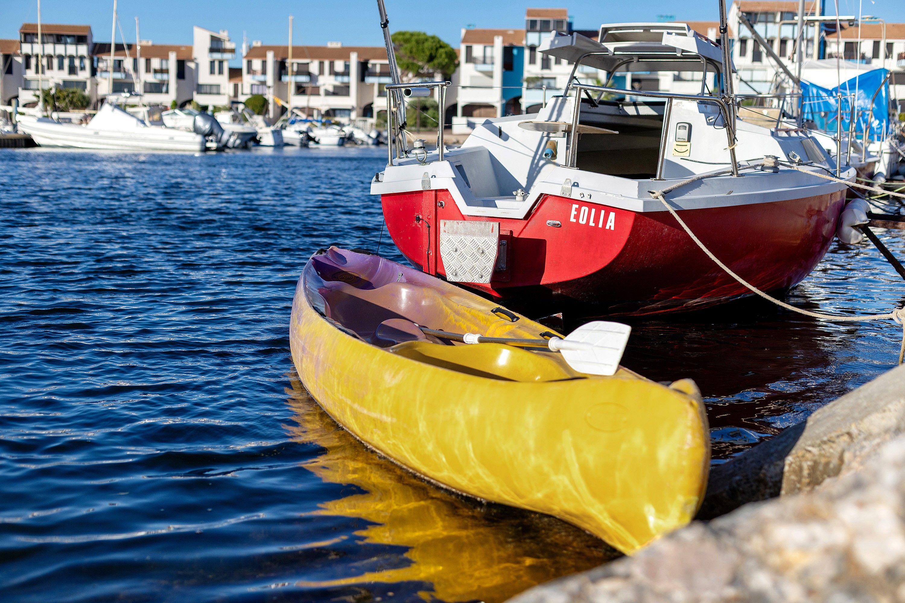 Een kajak vastgemaakt aan een rode boot met de naam 'EOLIA' in een jachthaven, met gebouwen op de achtergrond.