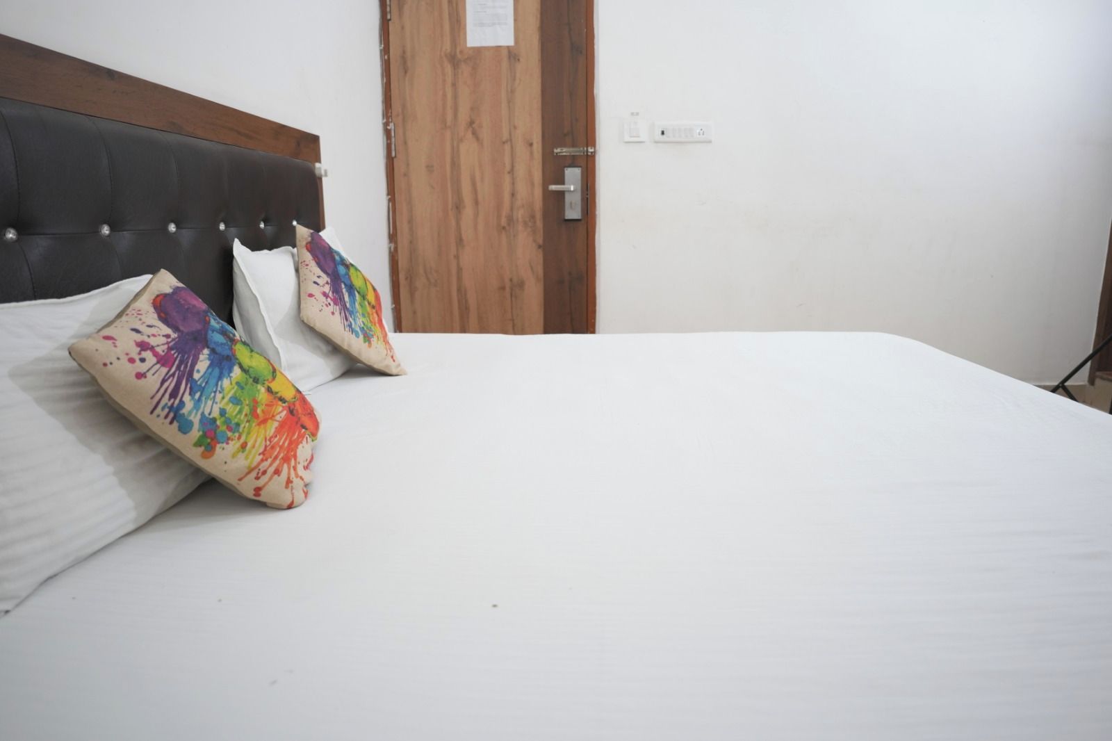 Una cama con sábana blanca y dos almohadas de colores, una puerta de madera y un interruptor de luz en la pared.