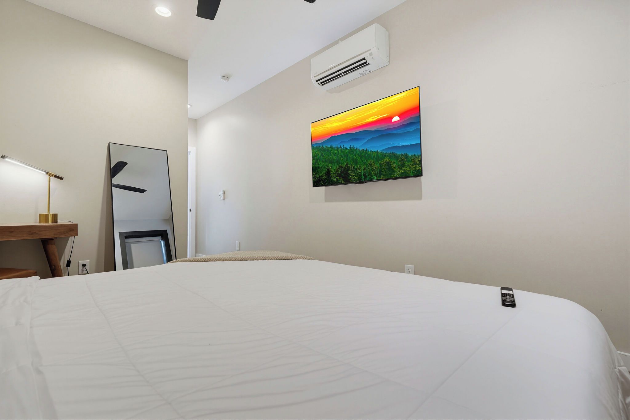 A hotel room with a large bed, a desk with a lamp, an air conditioner, and a painting of a sunset.