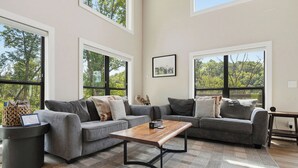 A spacious living room with a grey sectional sofa, a wooden coffee table, and large windows offering a view of trees. - Indoor Pool, Hot Tub & Mountain Views Near Pigeon Forge (Sevierville)