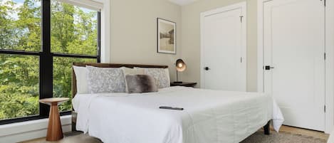 Un dormitorio con un ventanal grande, una cama con ropa de cama blanca, una lámpara de noche y un cuadro en la pared.