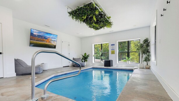 En inomhuspool med kristallklart blått vatten, ett grått loungeområde med en grå soffa och ett stort fönster som ger utsikt över omgivningen.
