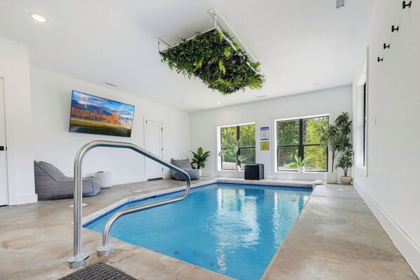 An indoor swimming pool with a clear blue water, a gray lounge area with a gray sofa, and a large window providing a view of the outdoors. - Indoor Pool, Hot Tub & Mountain Views Near Pigeon Forge (Sevierville)