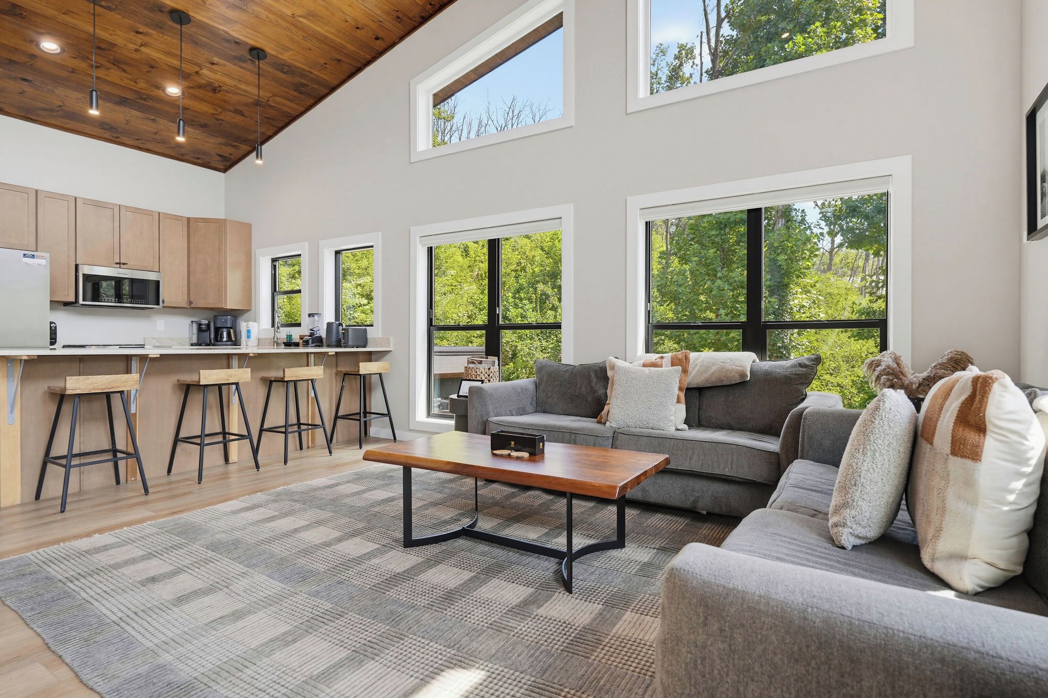 A modern living room with a sofa, coffee table, and bar stools. There is a kitchen area with a microwave and a large window.
