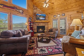 A cozy cabin interior with a fireplace, leather sofas, a television, and a large window overlooking a scenic landscape. - Smoky Mountain Views, Hot Tub & Game Loft Retreat (PIGEON FORGE)