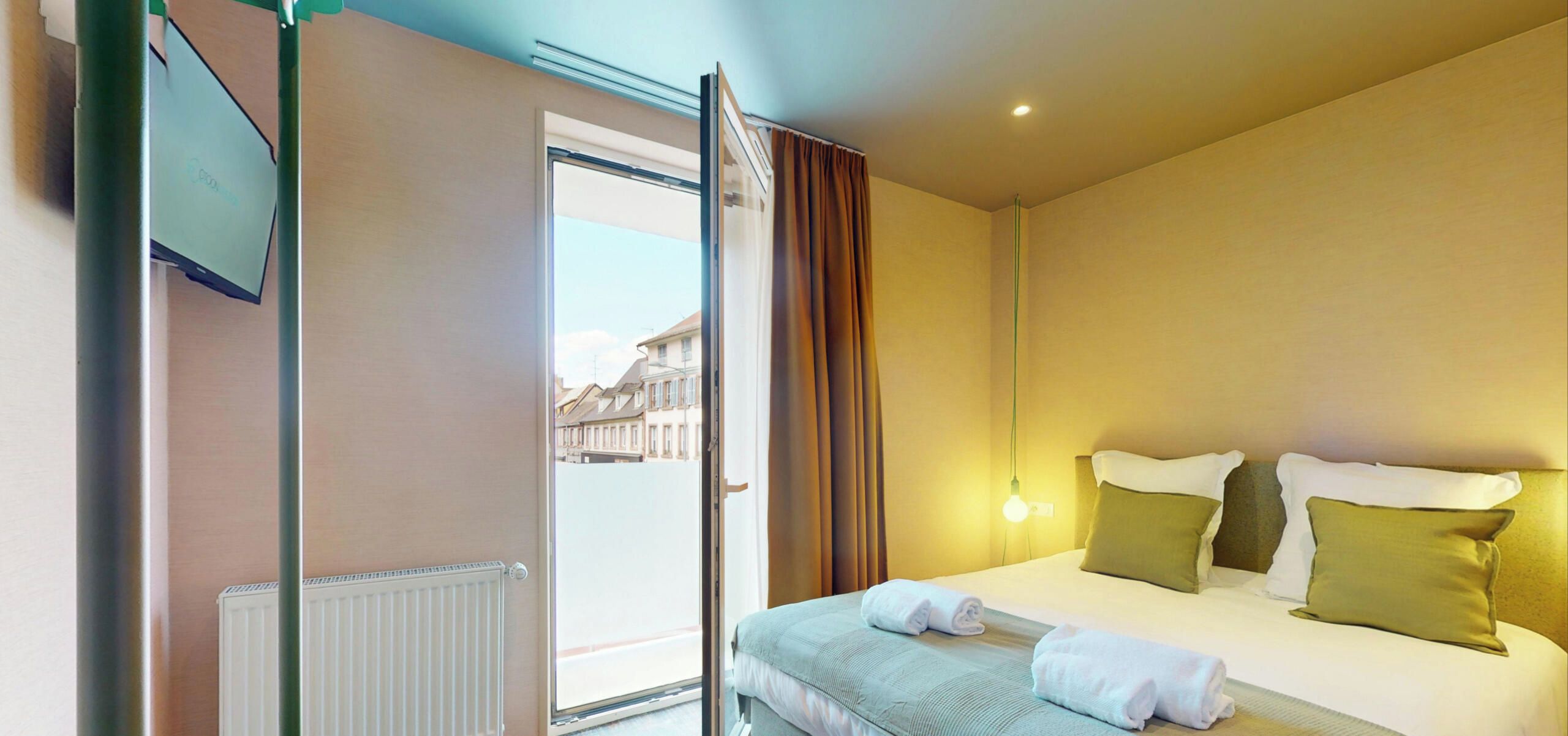 Quarto de hotel com cama, radiador, janela com cortinas e vista para edifícios.