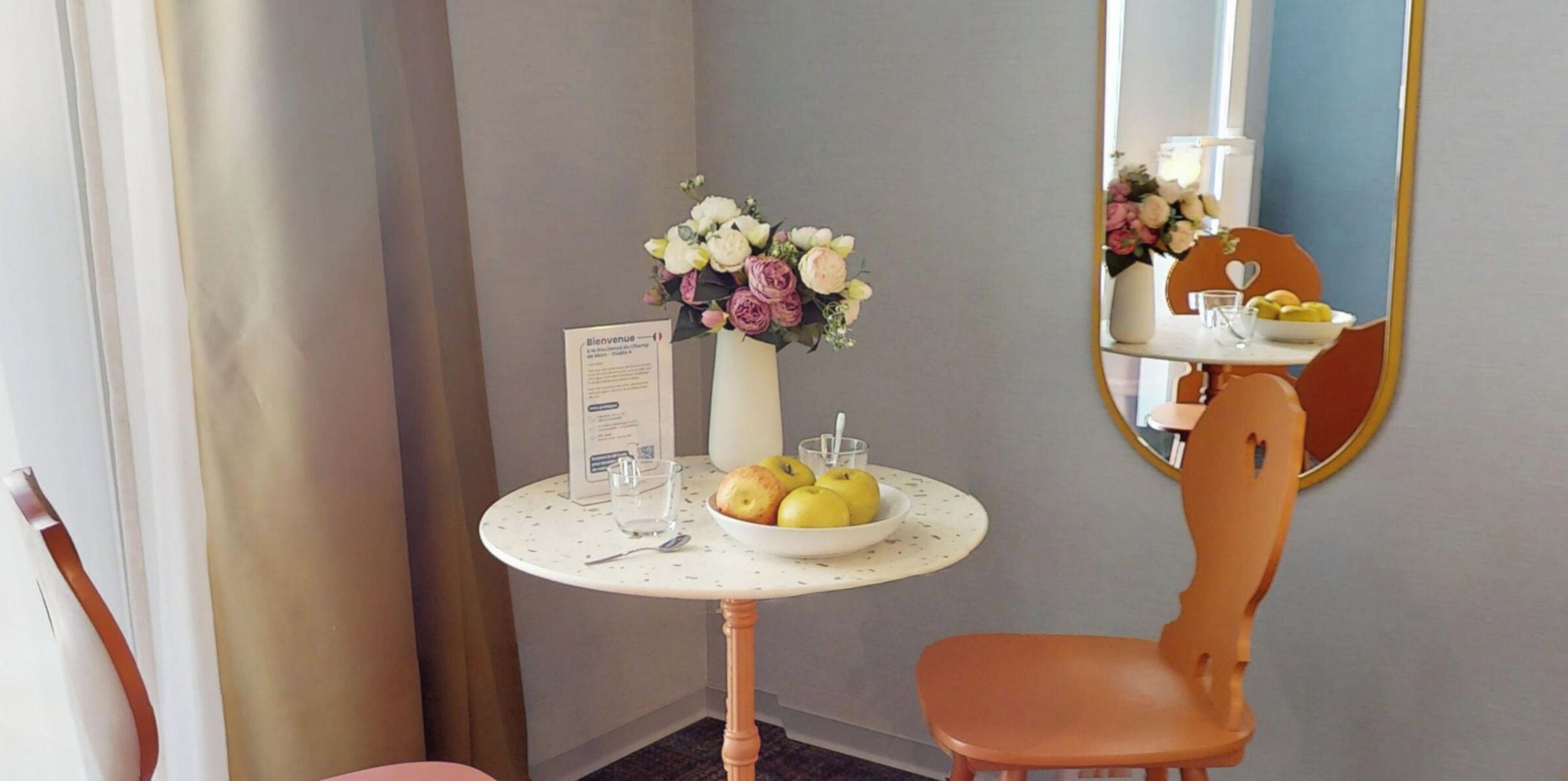 A hotel room with a round table, a vase of flowers, a bowl of fruit, a chair, and a mirror.
