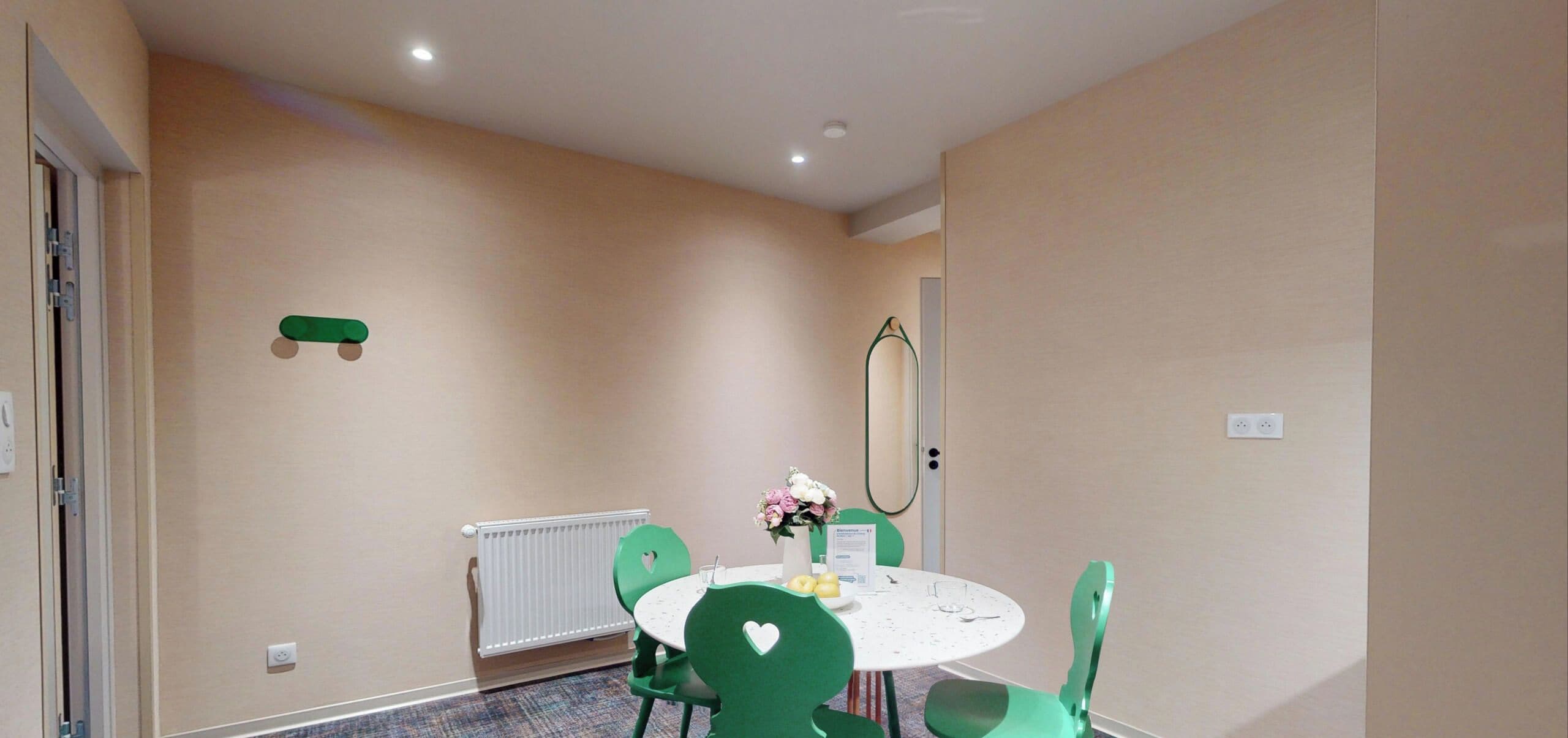 A room with a round table, green chairs, a radiator, a mirror, and a door.