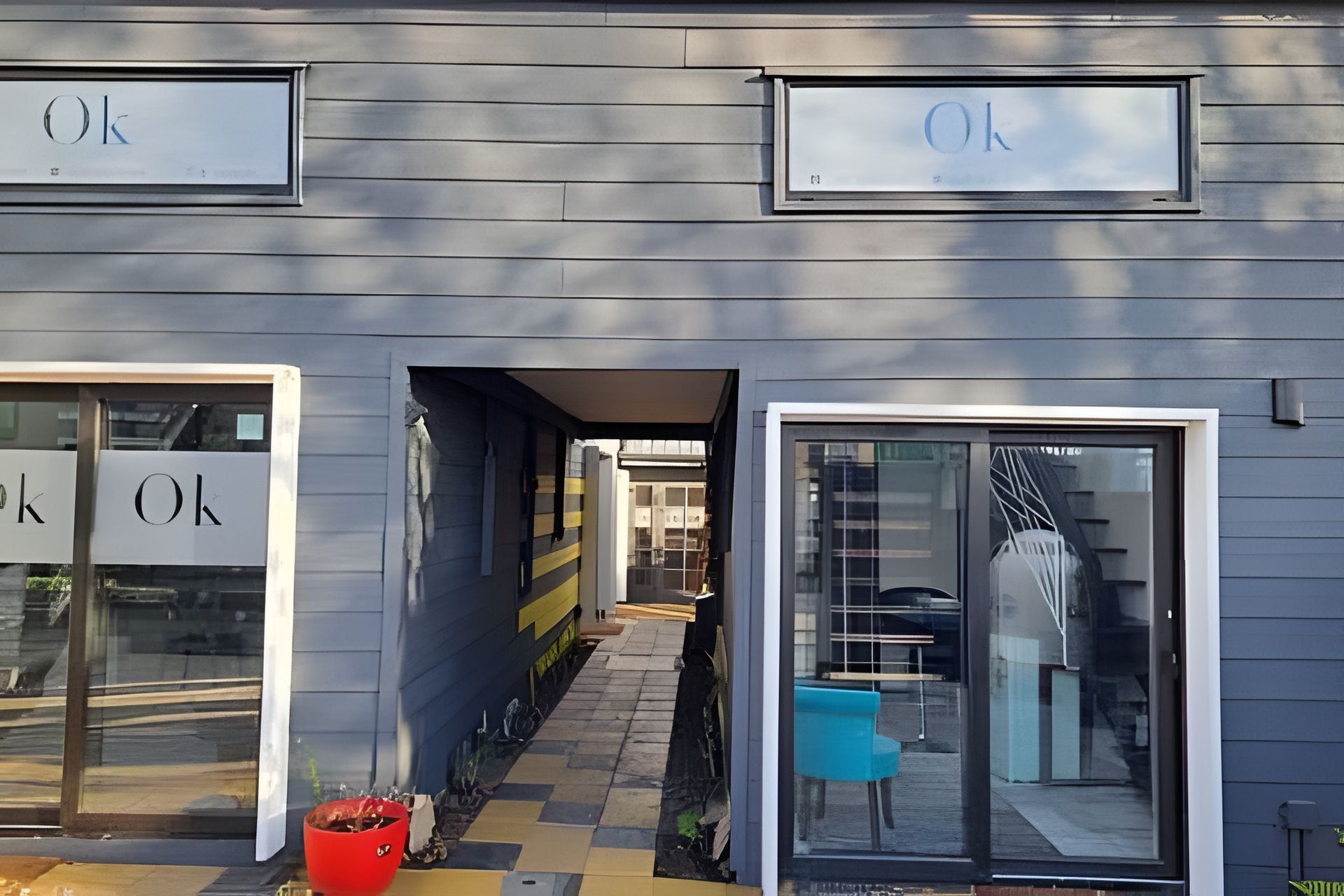 Fachada de edificio con letreros que dicen 'Ok' sobre las puertas de entrada, un sendero que conduce a una zona techada, y una silla azul afuera.