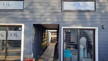 Fachada de edificio con letreros que dicen 'Ok' sobre las puertas de entrada, un sendero que conduce a una zona techada, y una silla azul afuera.