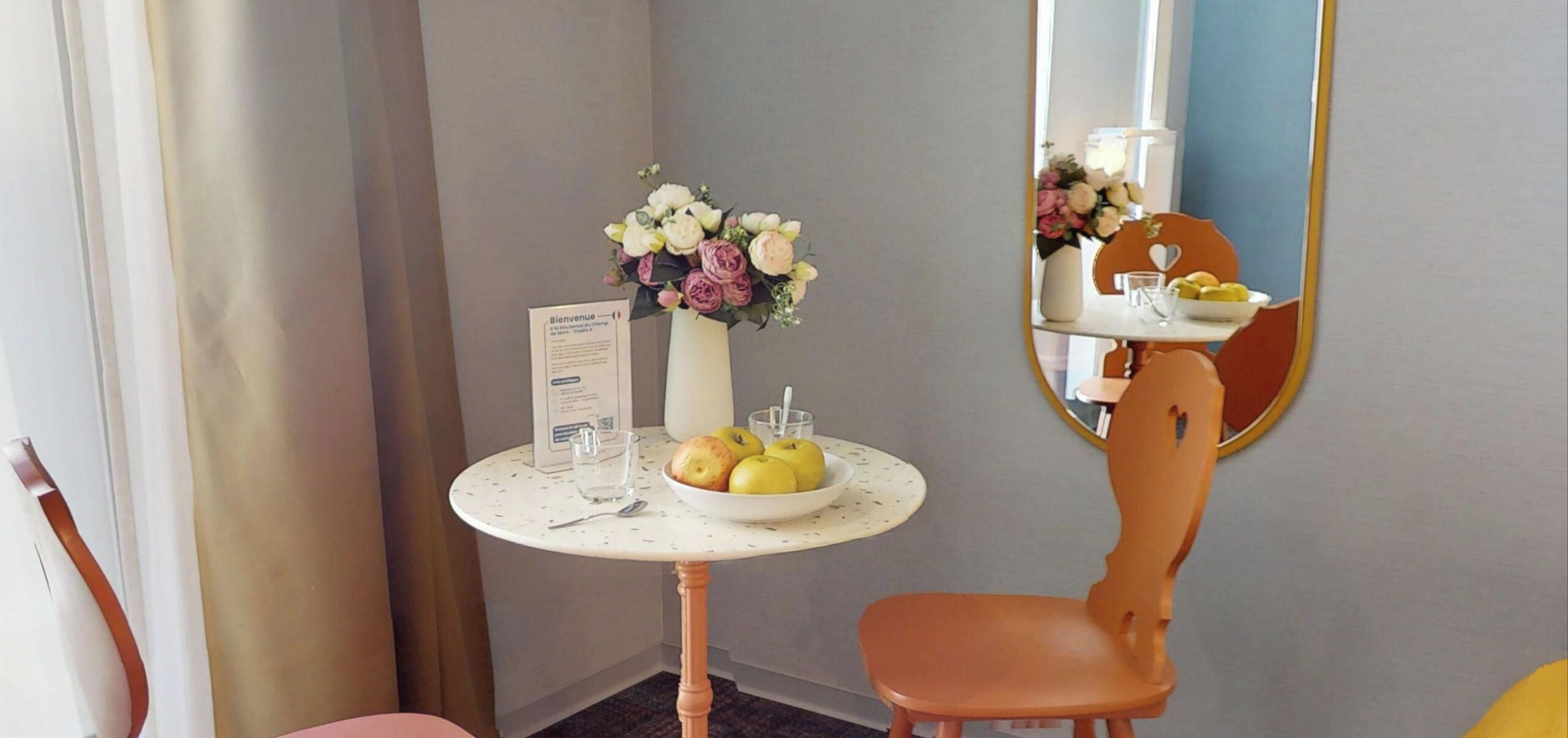 Ein Hotelzimmer mit einem runden Tisch, einer Blumenvase, einer Obstschale, einem Sessel und einem Spiegel.