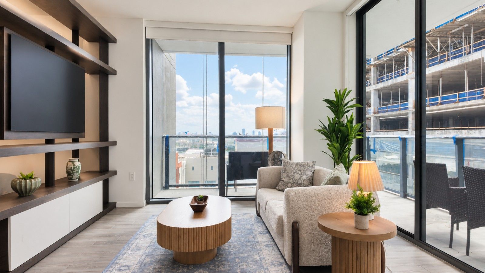 A modern living room with a flat-screen TV, a sofa, a coffee table, and a balcony with a view of a construction site.
