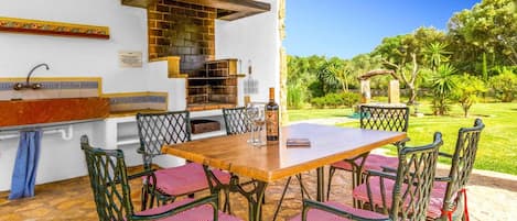An outdoor patio with a wooden table and chairs, a brick oven, and a view of a garden.