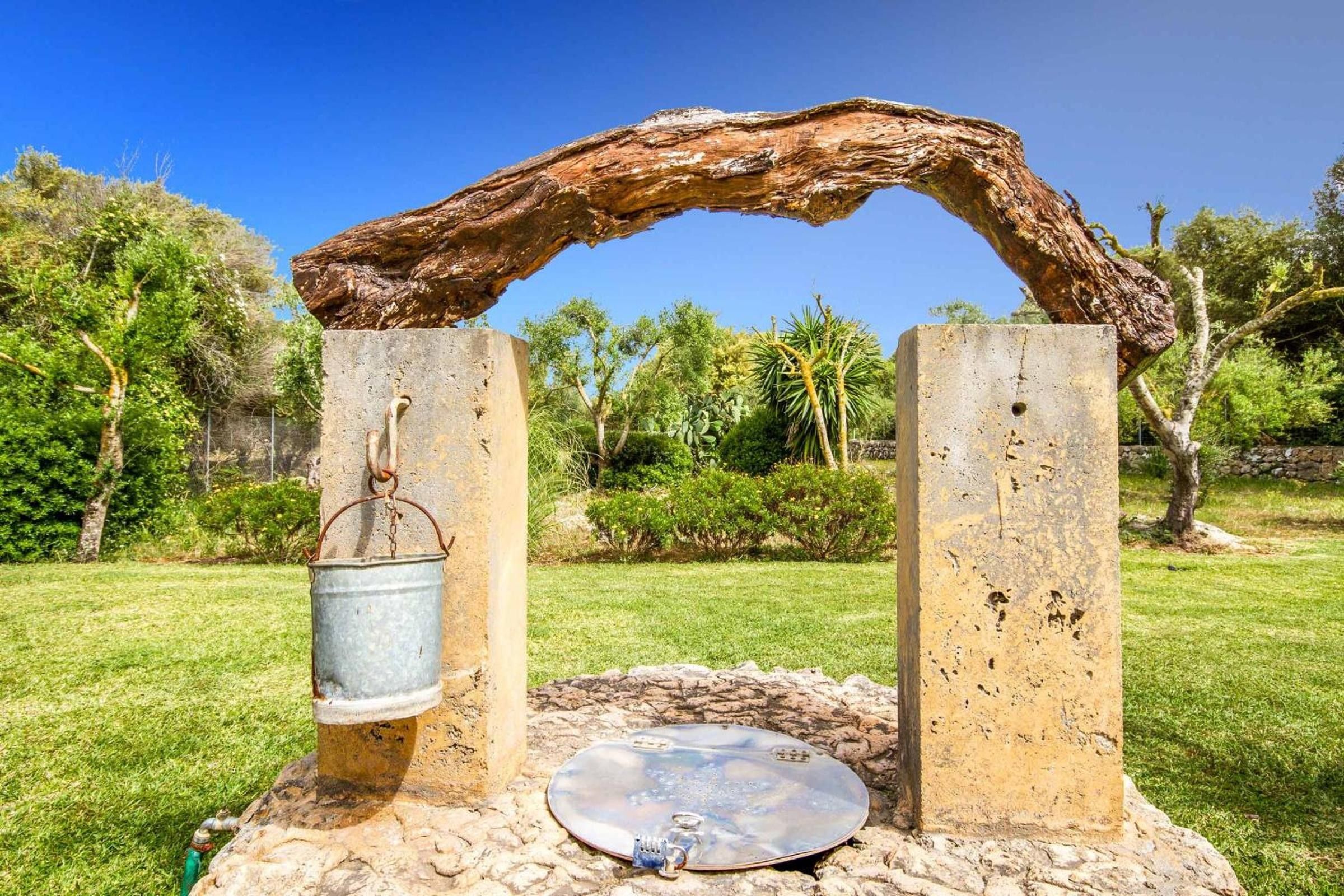 A rustic outdoor structure with a metal bucket and a flat surface, possibly for seating or decoration, set against a backdrop of greenery and trees.