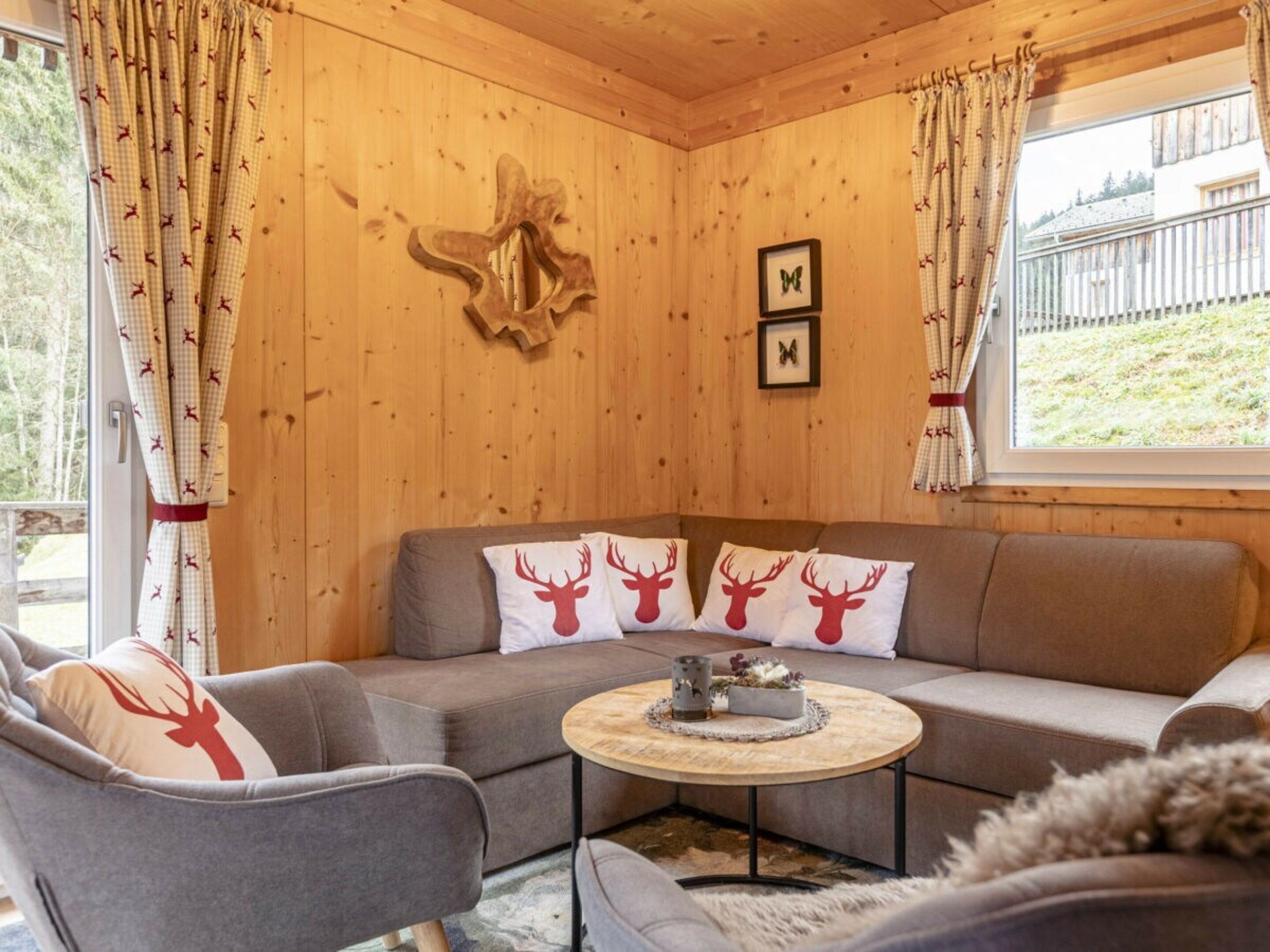 A cozy room with a wooden interior, a sofa with reindeer-themed cushions, a round coffee table, and a window with curtains.