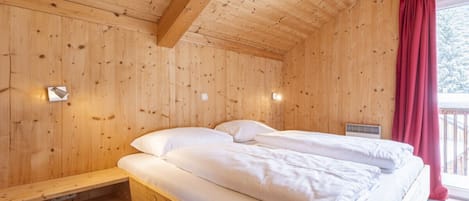 A wooden cabin room with a bed, a window with red curtains, and a small shelf.
