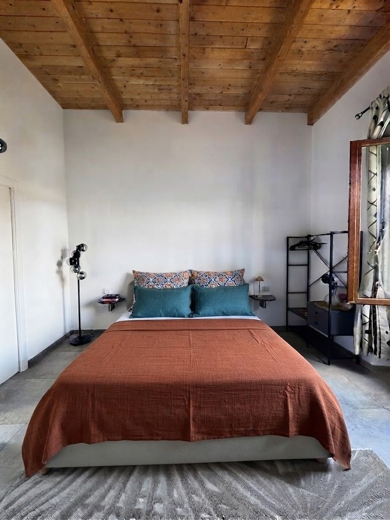 Una camera da letto con soffitto in legno, un letto con coperta marrone, due cuscini, una lampada da terra e una mensola metallica con diversi oggetti.
