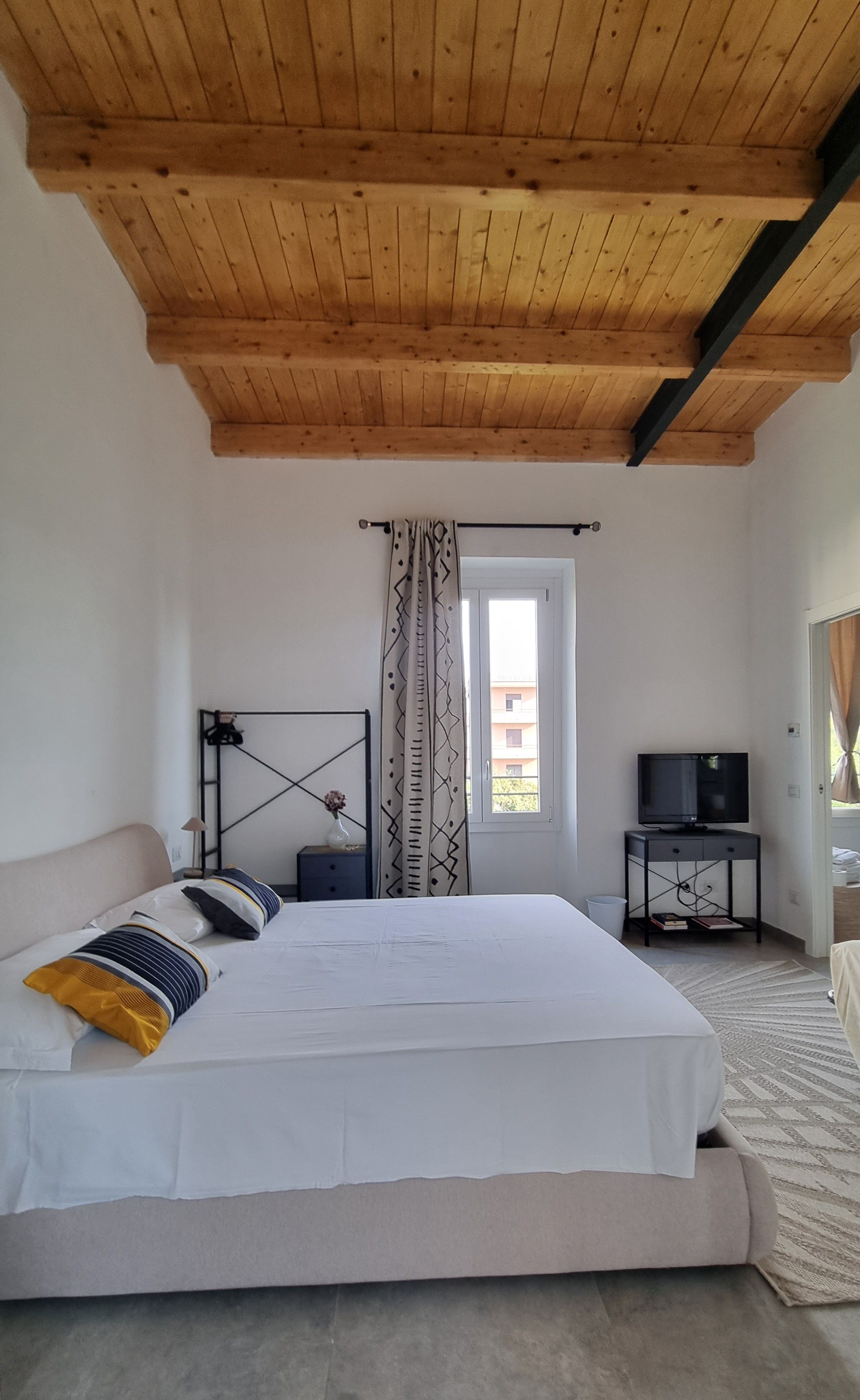 A bedroom with a wooden ceiling, a bed with white bedding, a TV on a stand, and a window with curtains.