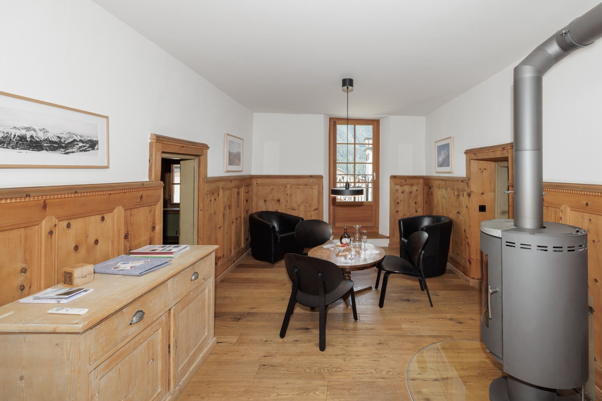 Een kamer met houten lambrisering, een eettafel met stoelen, een zwarte leren bank, een houten kast en een houtkachel.