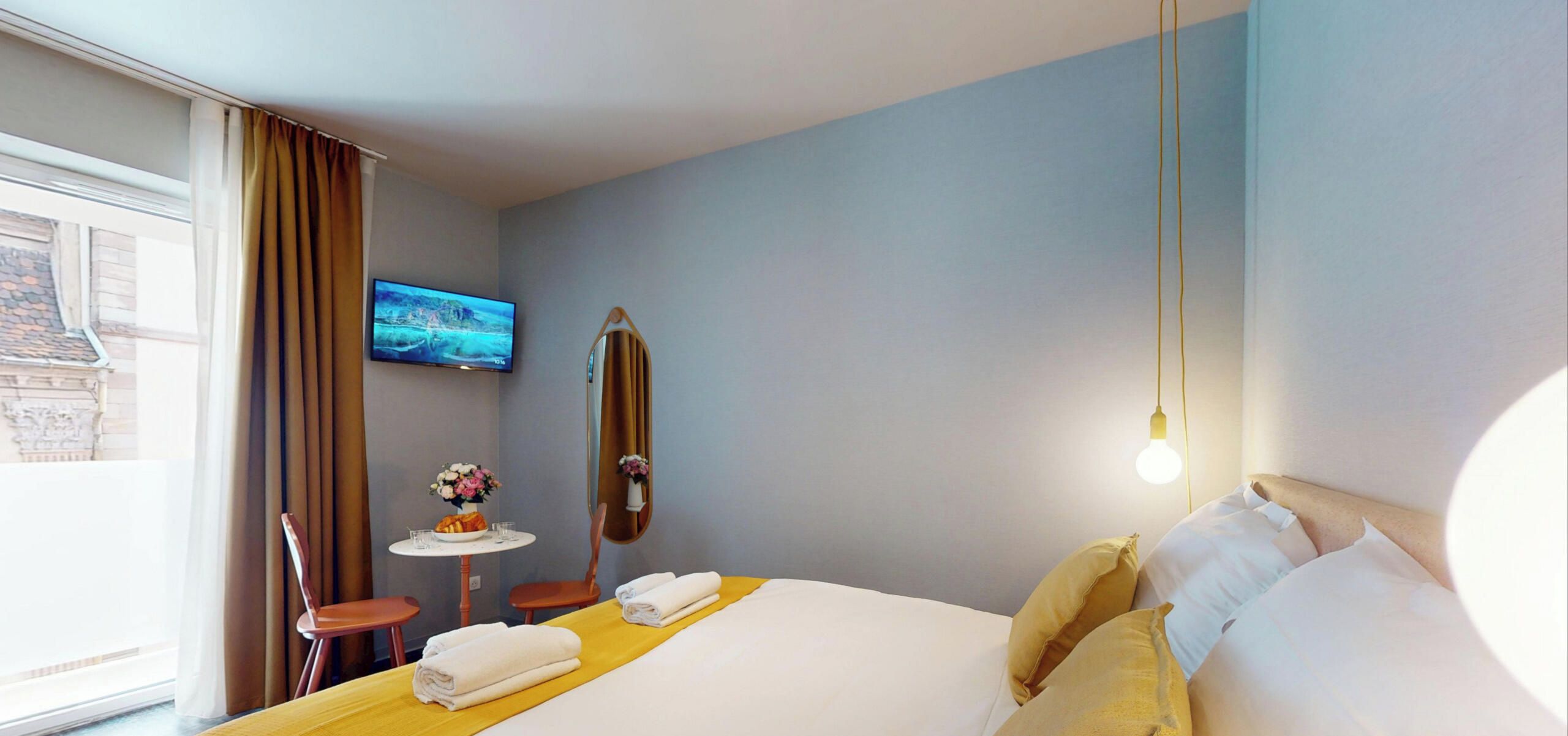 A hotel room with a bed, a small table with a vase of flowers, a chair, a television on the wall, and a window with curtains.