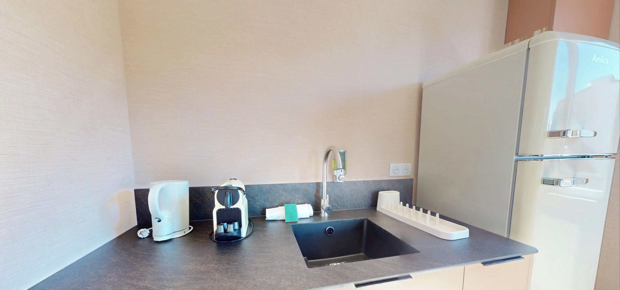 Eine kleine Küchenzeile mit Spüle, Kaffeemaschine, Wasserkocher und Kühlschrank.