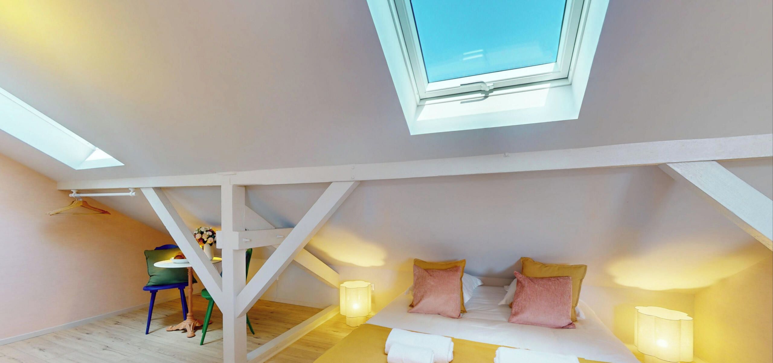 Ein Zimmer mit schrägverdeckter Decke, einem Bett mit Kissen, einem Tisch mit Stühlen und einem Oberlicht.