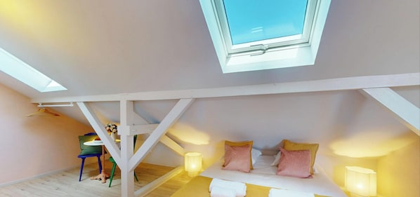 Una habitación con techo inclinado, una cama con almohadas, una mesa con sillas y un tragaluz.