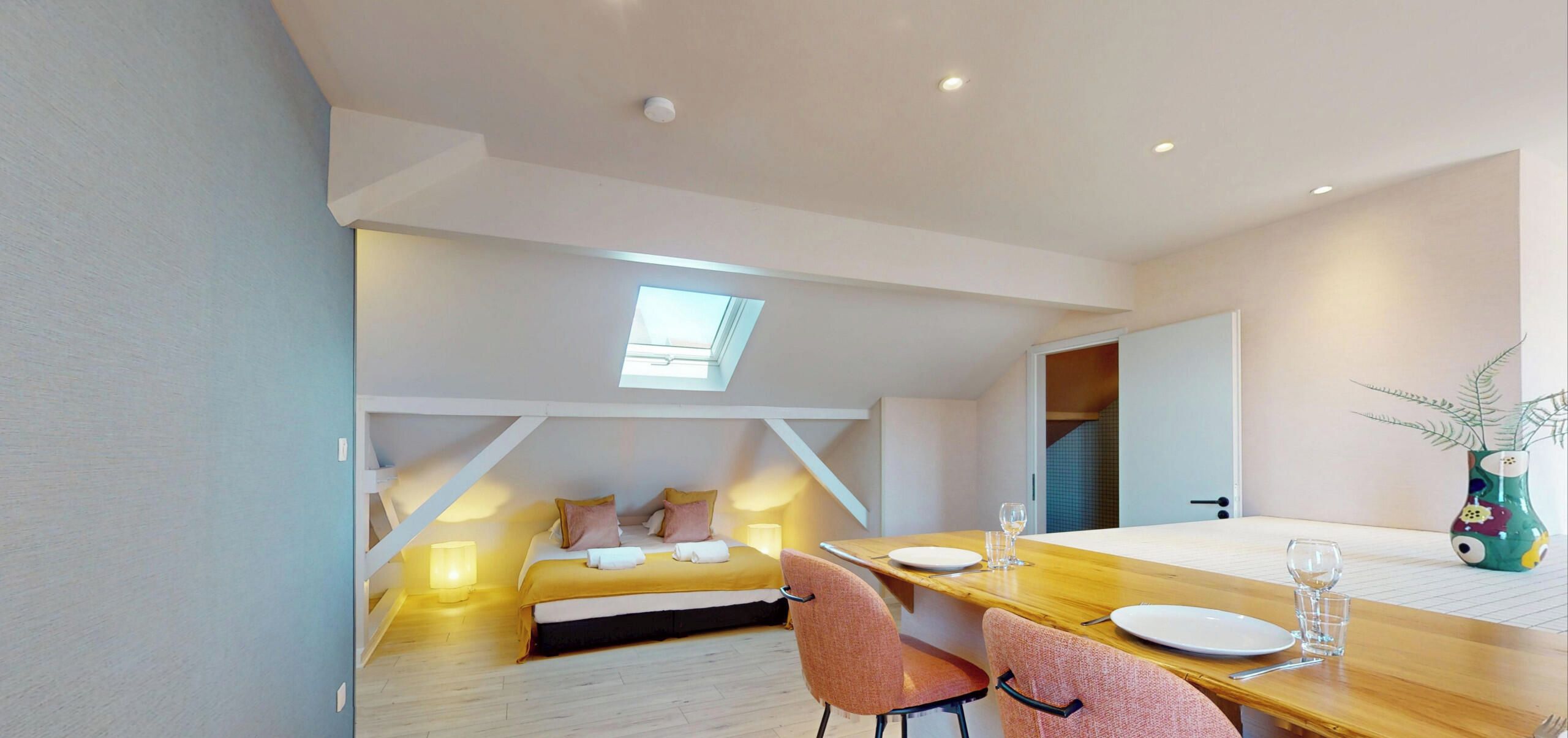 A modern bedroom with a skylight, a wooden dining table set for two, and a view into the adjacent room.