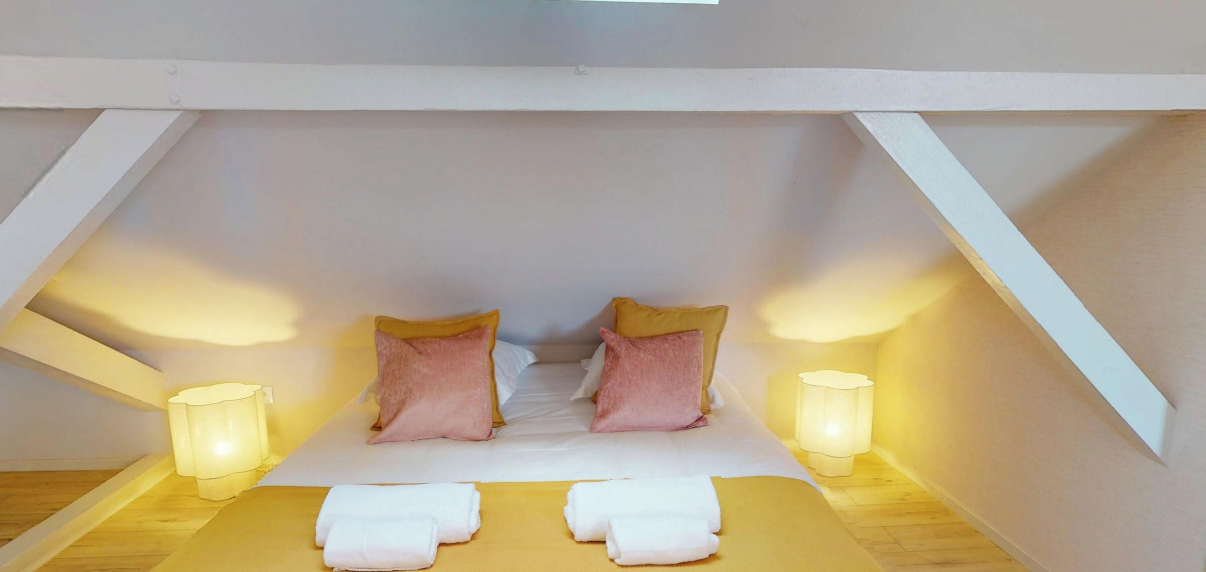 A small, cozy room with a sloped ceiling, two bedside lamps, and two pillows.