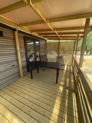 An outdoor wooden deck with a table and chairs, a covered area with a roof, and a view of trees in the background. - Camping les Chênes Mobile Home (new) in Porto Vecchio, 50 m from the sea (Porto-Vecchio)