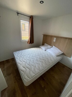 A single bed with a headboard, a window with curtains, and a light switch on the wall. - Camping les Chênes Mobile Home (new) in Porto Vecchio, 50 m from the sea (Porto-Vecchio)