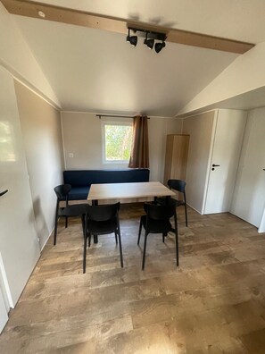 A room with a wooden dining table, four chairs, a sofa, and a window with curtains. - Camping les Chênes Mobile Home (new) in Porto Vecchio, 50 m from the sea (Porto-Vecchio)