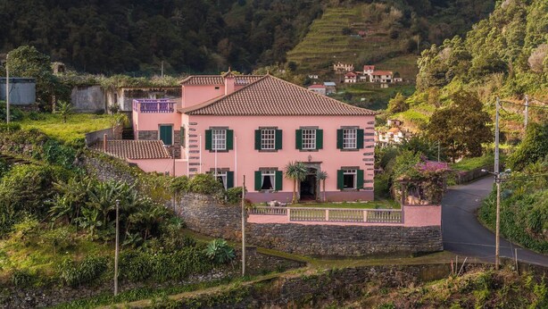 Eine pinkfarbene Villa mit Ziegeldach, umgeben von üppigem Grün und einer Steinmauer.