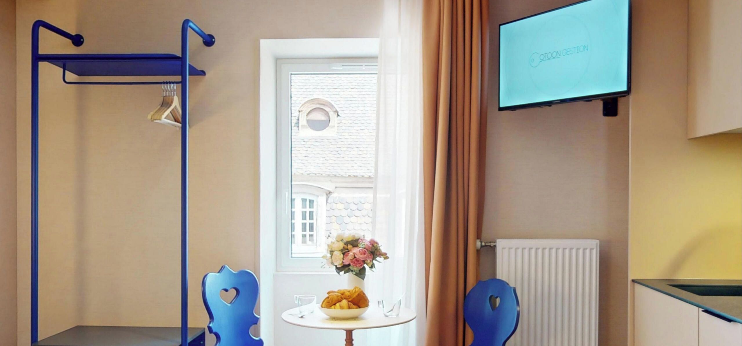Ein Hotelzimmer mit Bett, Wandfernseher, Fenster mit Vorhängen und einem Tisch mit Blumen.'