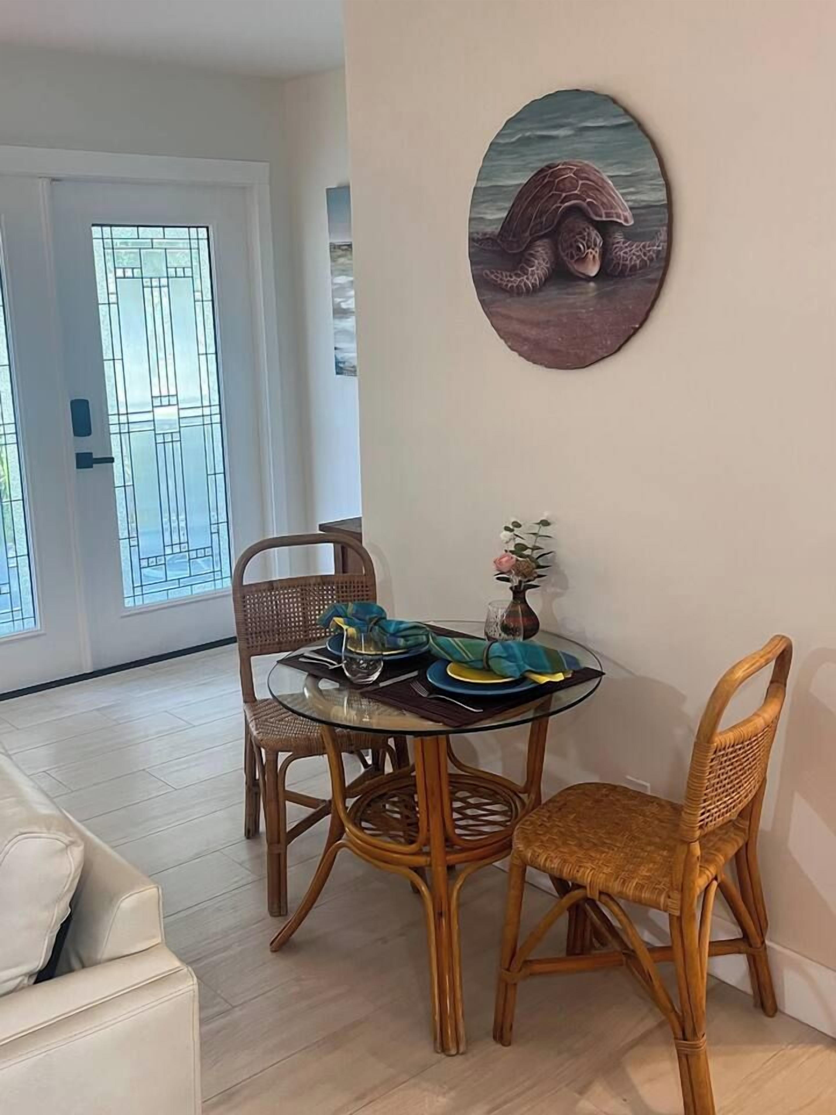 A dining area with a round glass table, wicker chairs, and a wall-mounted turtle artwork.