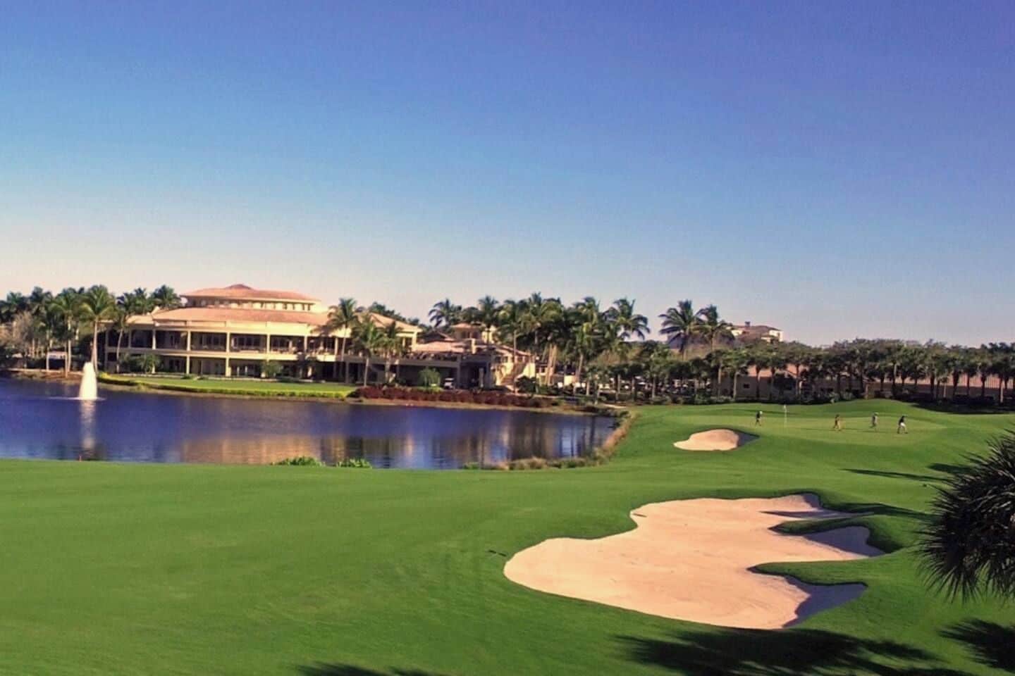 A golf course with a pond, palm trees, and a building in the background.