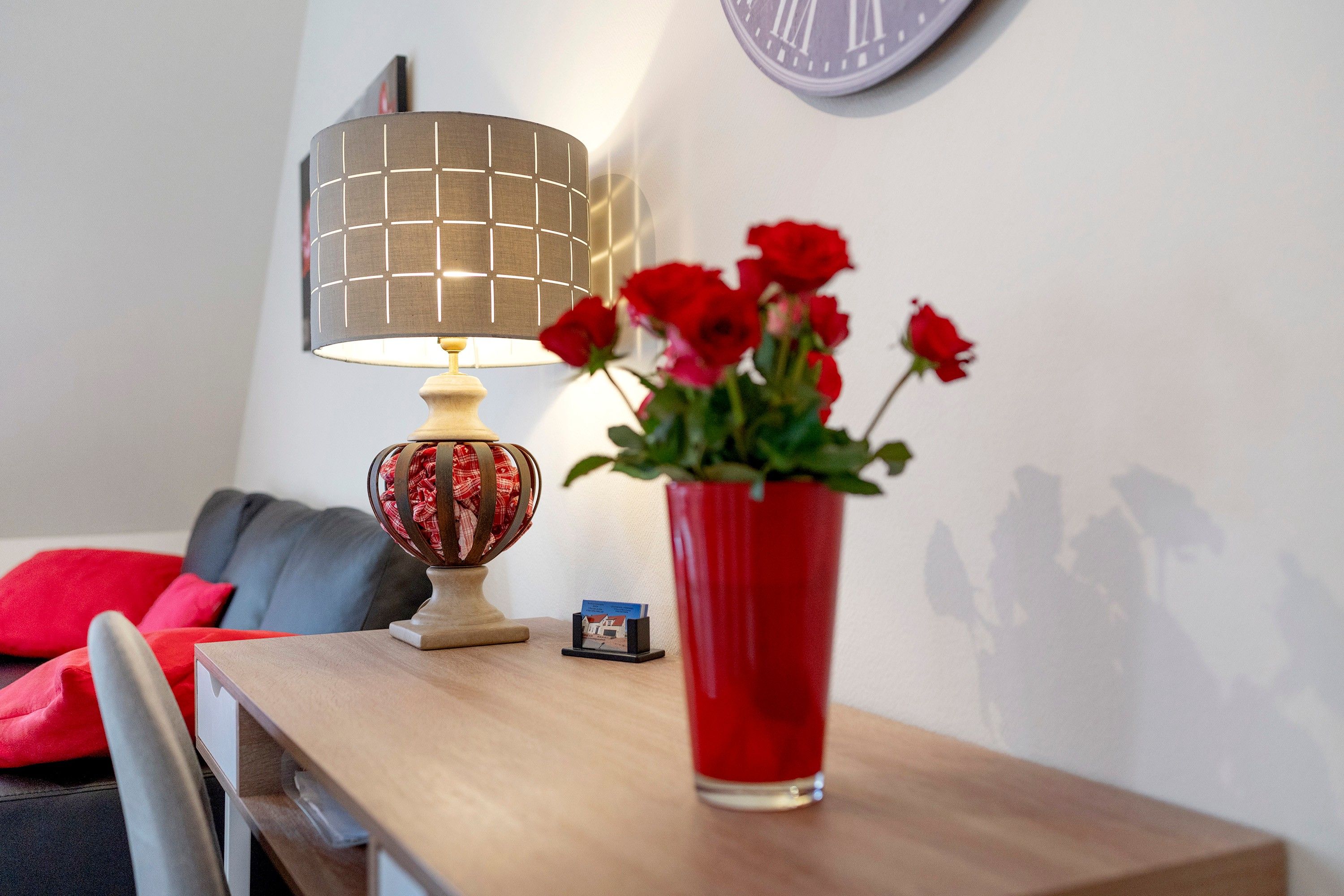 A wooden table with a red vase of flowers, a lamp with a grid-patterned lampshade, and a small card stand with a brochure.