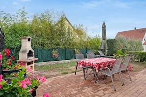 Une terrasse avec un sol en briques, une table recouverte d’une nappe rouge, des chaises en métal, un four en pierre et une clôture verte.