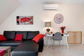 A modern living room with a black leather sofa, red cushions, a desk with a chair, a wall clock, and an air conditioning unit.
